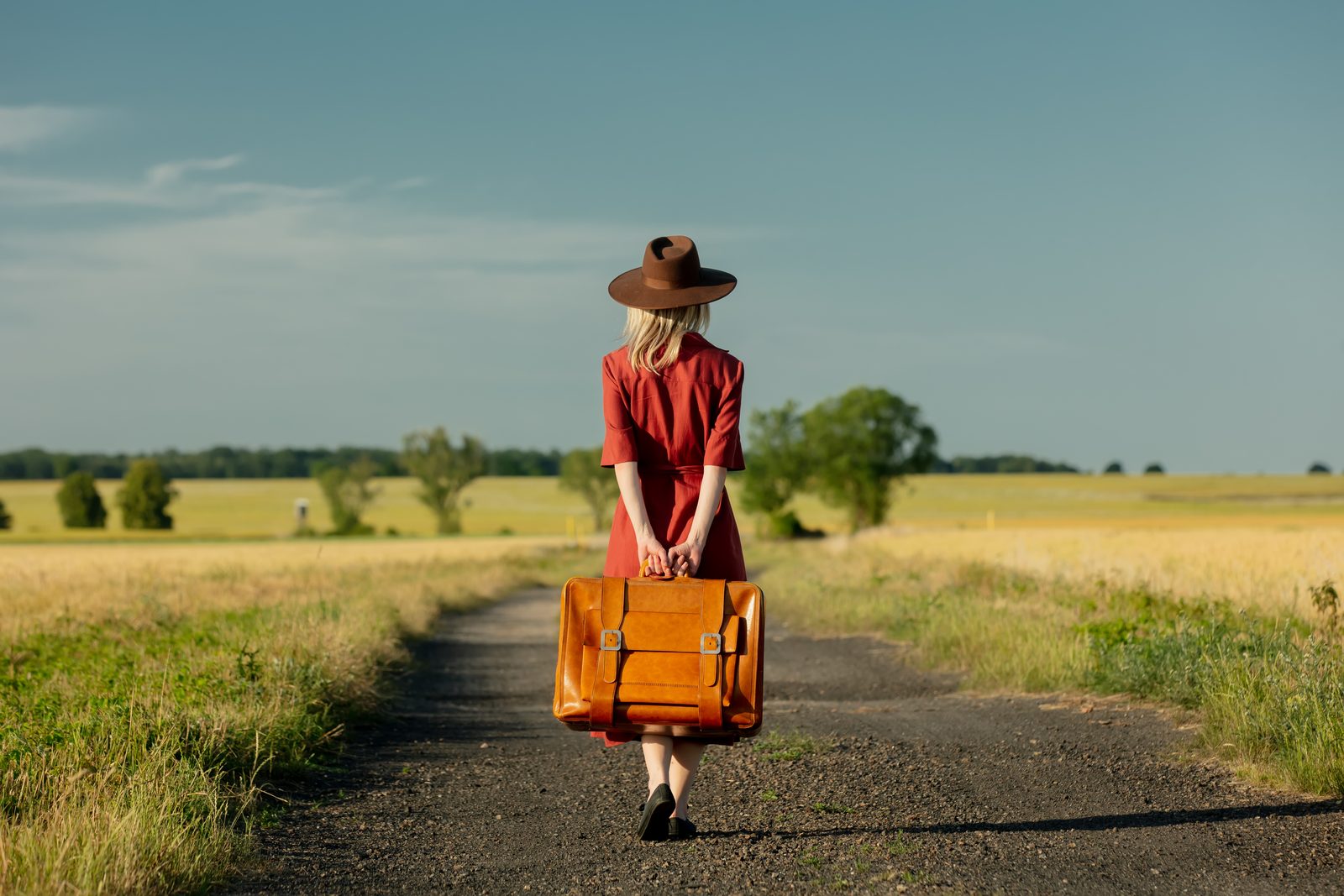 A traveler in a red dress and hat walking down a country dirt road with a vintage leather suitcase